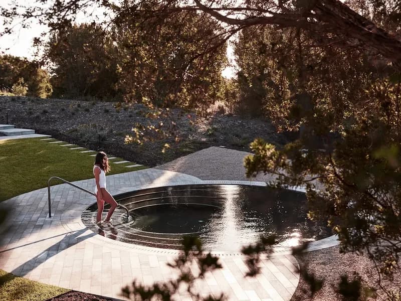 A bather stepping into a round open-air thermal pool under the trees, in late sun