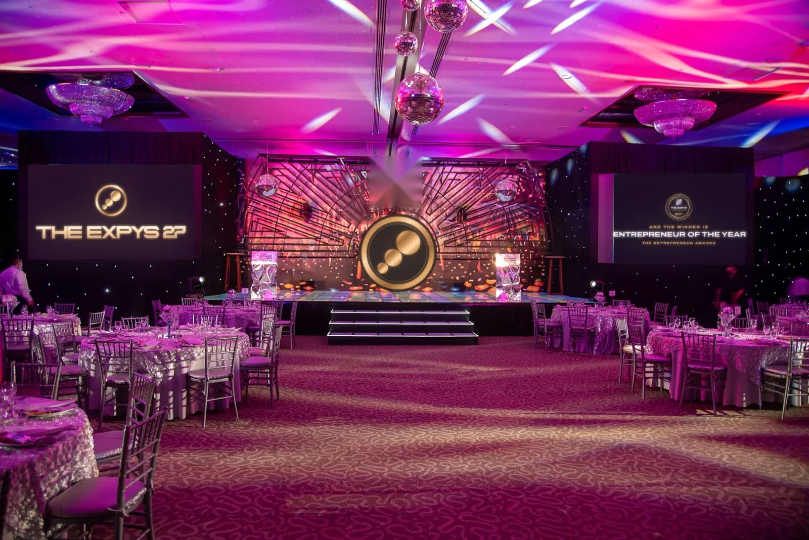 A violet-lit ballroom with a stage between two screens, THE EXPYS 27 on the left screen