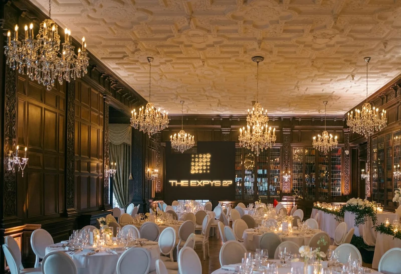 A panelled library ballroom under crystal chandeliers, white rounds of ten, THE EXPYS 27 on the screen at the far end