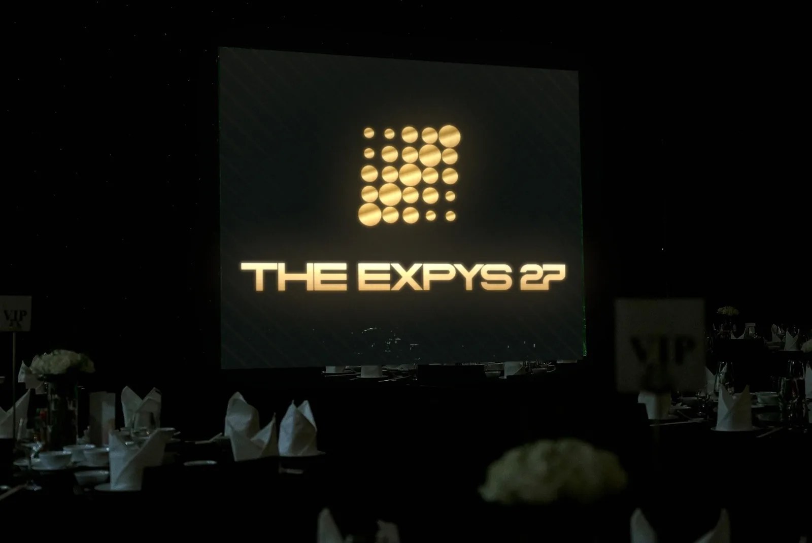 A dark room of dressed rounds, THE EXPYS 27 on one big screen over the tables