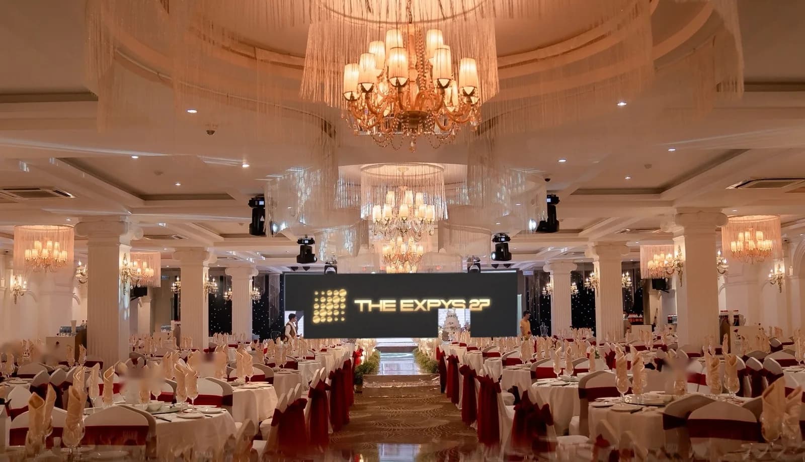 A white-columned ballroom under fringed chandeliers, THE EXPYS 27 on the LED wall over the stage