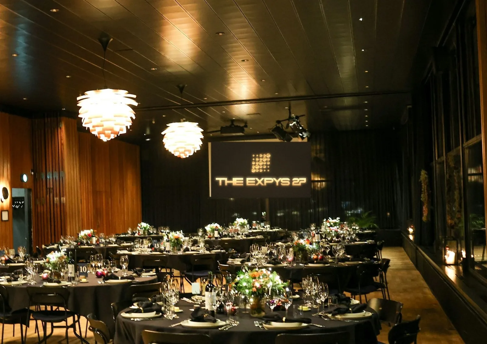 A timber-panelled hall set with rounds of ten under pendant lamps, THE EXPYS 27 on the end-wall screen
