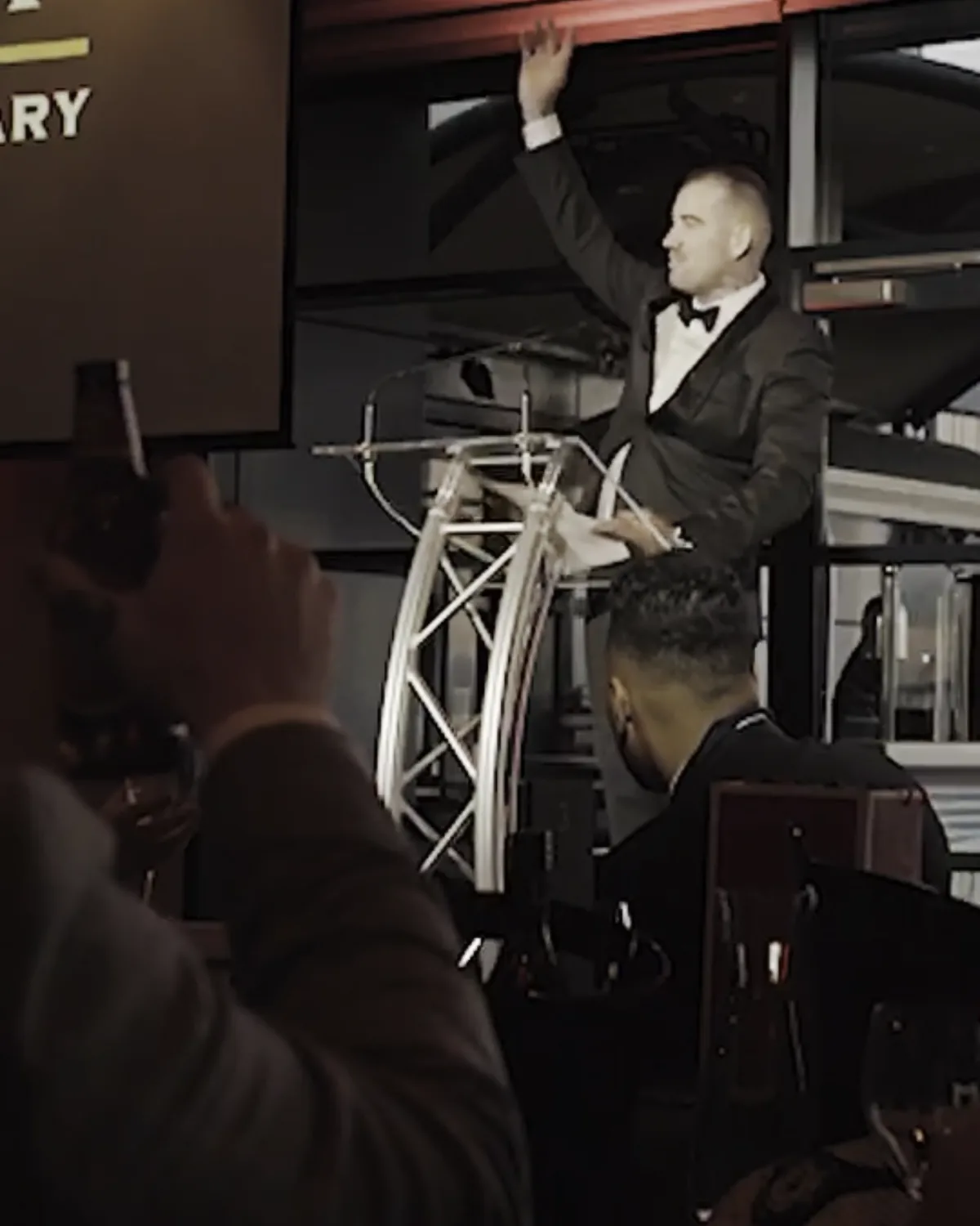 A host in black tie at a truss lectern, arm raised mid speech, the room watching from below