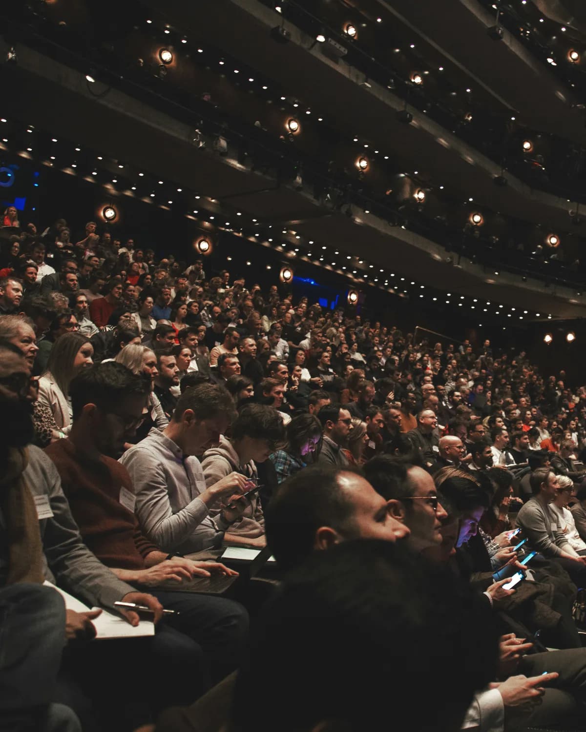 A packed conference plenary seen from the side, delegates banked up the tiers with notepads and phones, balconies full above them