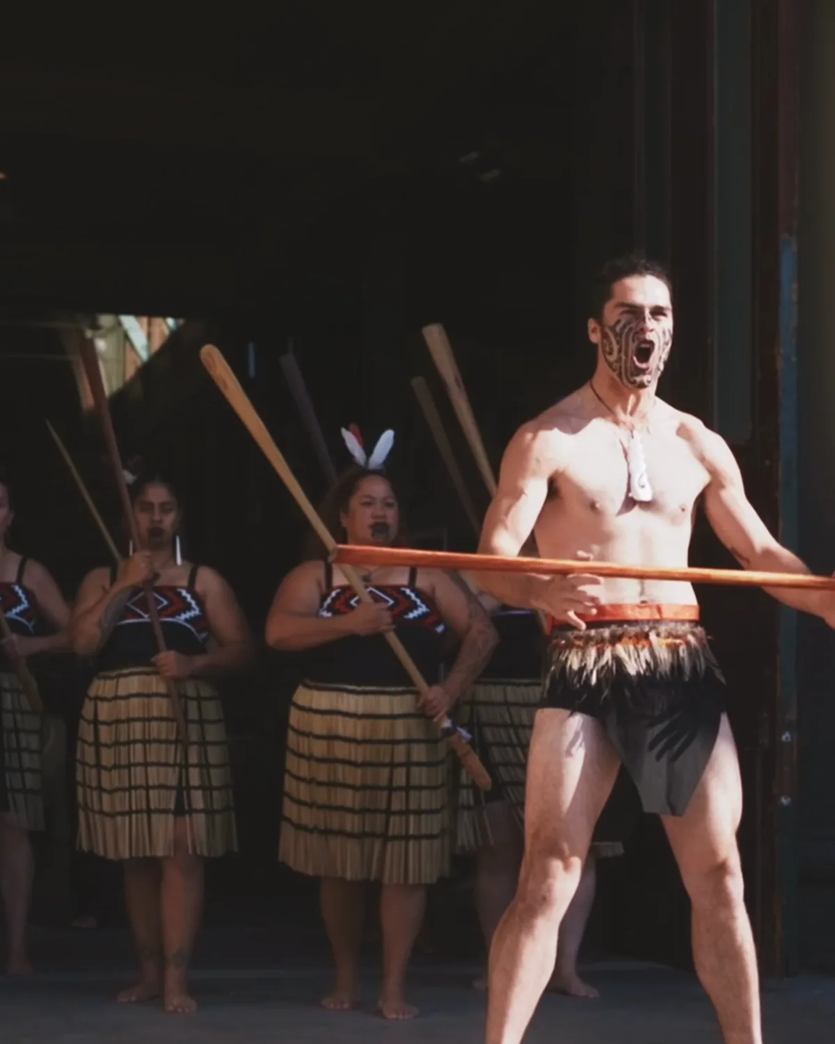 A haka performer mid challenge with a taiaha raised, his group standing behind him on the forecourt