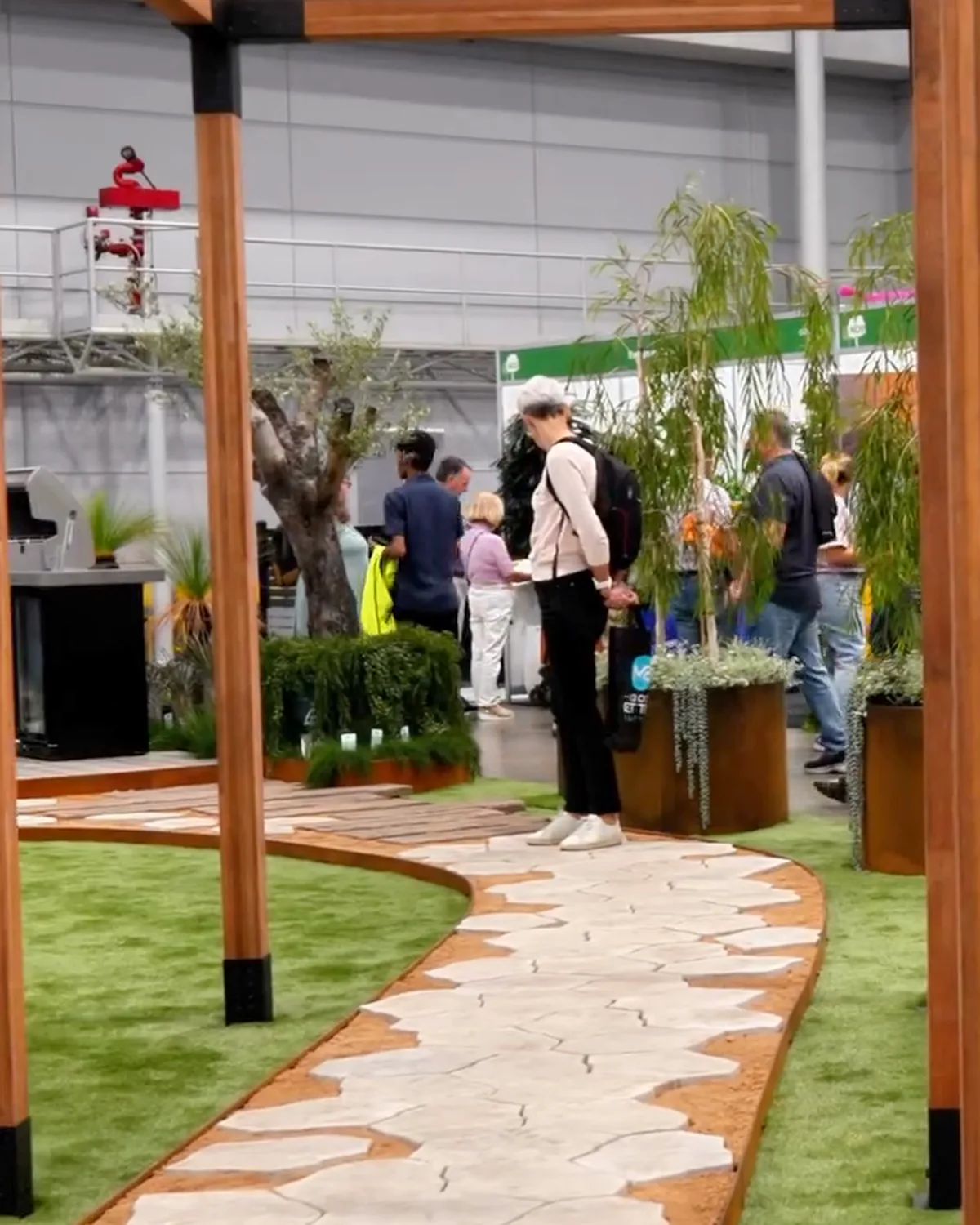 Visitors browsing a built landscape display on a home show floor, a timber pergola over crazy paving and turf, exhibitor stands down the hall behind