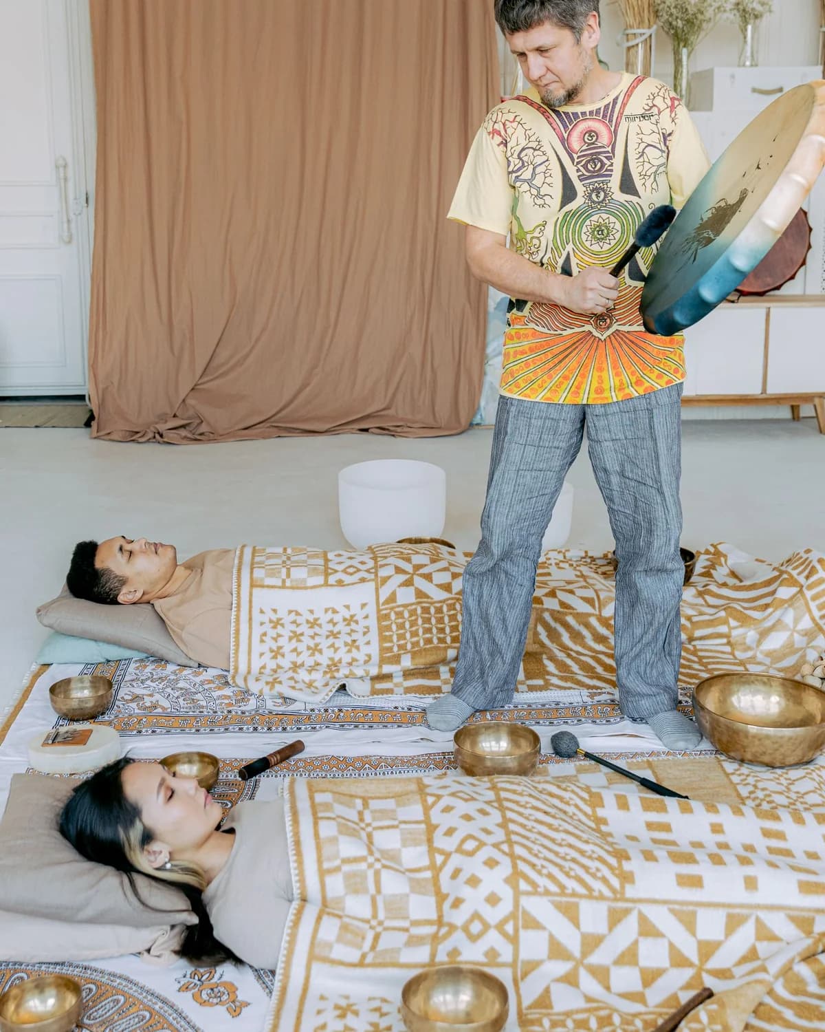 A sound practitioner over two people resting on mats, singing bowls set around them at a wellbeing session