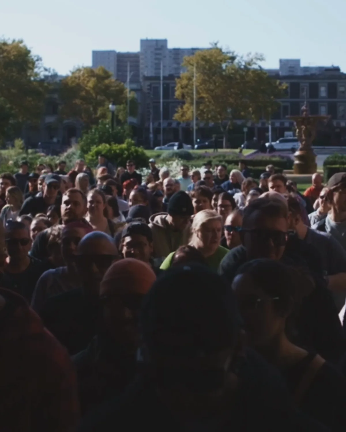 A daytime queue winding across the park towards the gates, the city skyline behind the trees