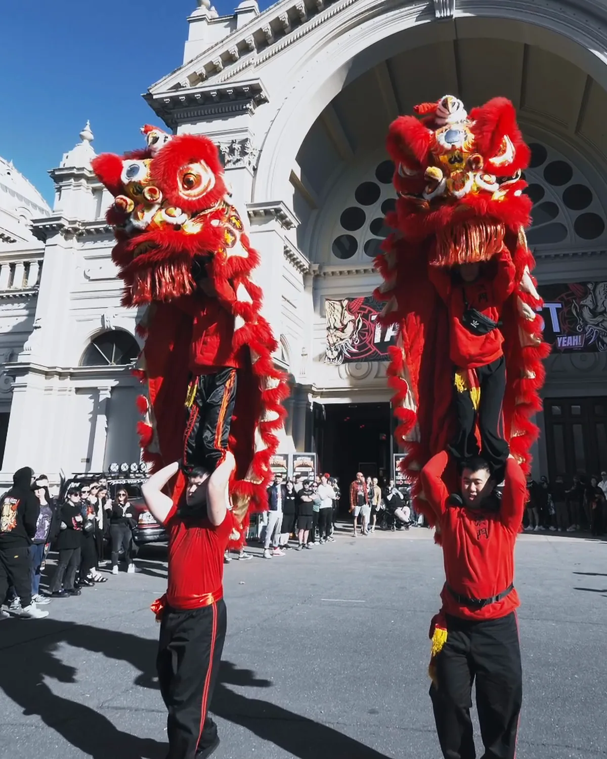 Two red lion dancers on stilts in front of the Royal Exhibition Building, a crowd lining the forecourt