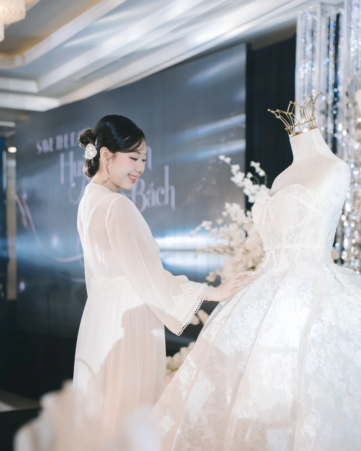 A visitor at a bridal expo reaching to a lace gown crowned on its mannequin, the show backdrop behind her