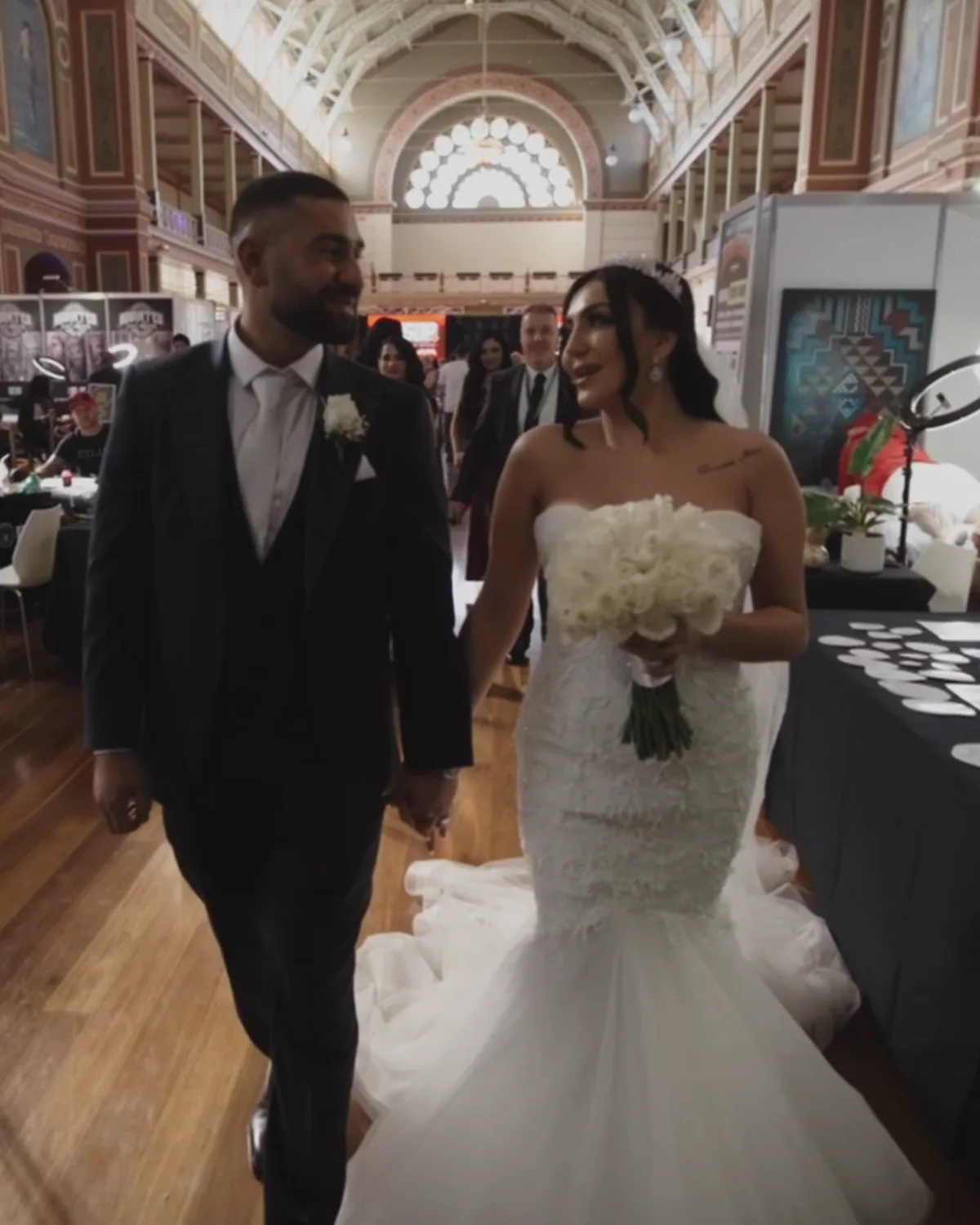 A bride and groom walking hand in hand down the aisle of a heritage exhibition hall, her train behind her